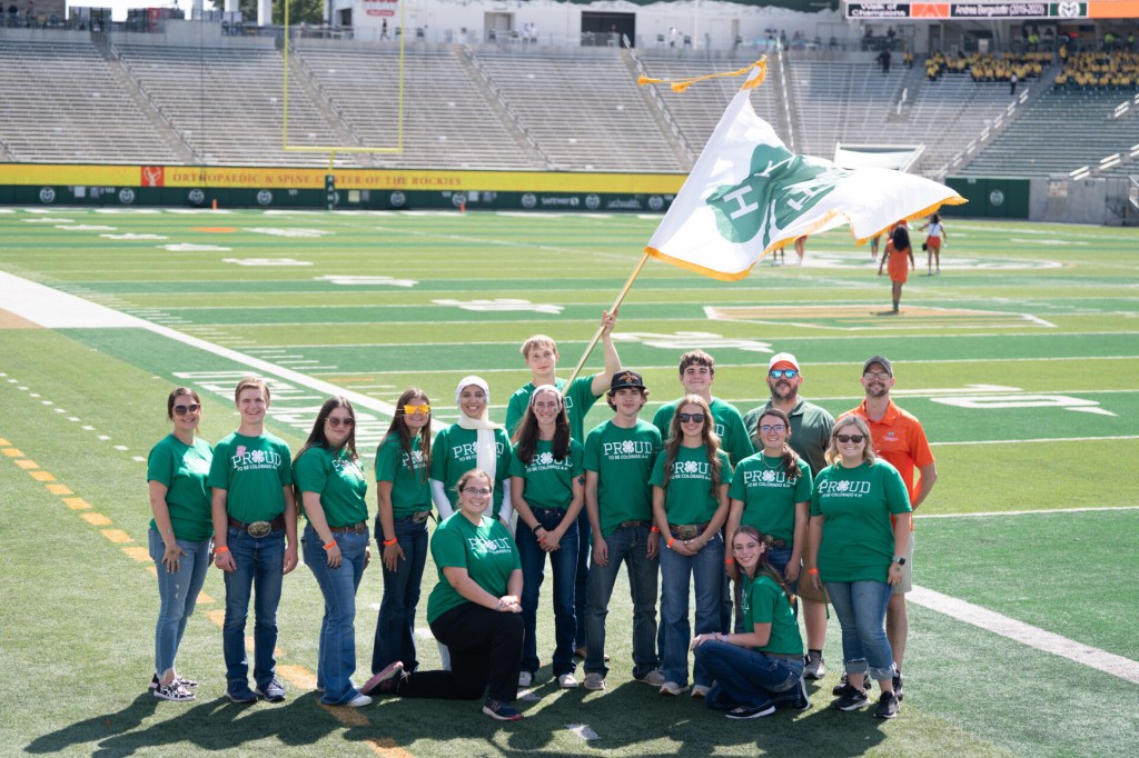 A group of 4-H youth and adults stand together on the field at Canvas Stadium, smiling for a group photo. One participant holds a large 4-H flag..