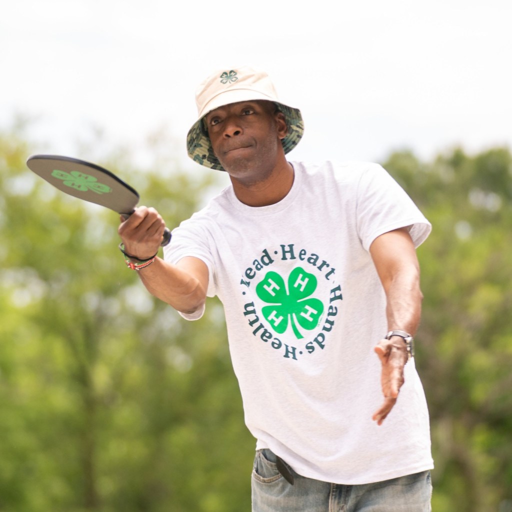 A man wearing a white 4-H T-shirt and a bucket hat plays pickleball outdoors, extending his arm forward as he hits the ball with a paddle. Trees fill the background on a bright day.
