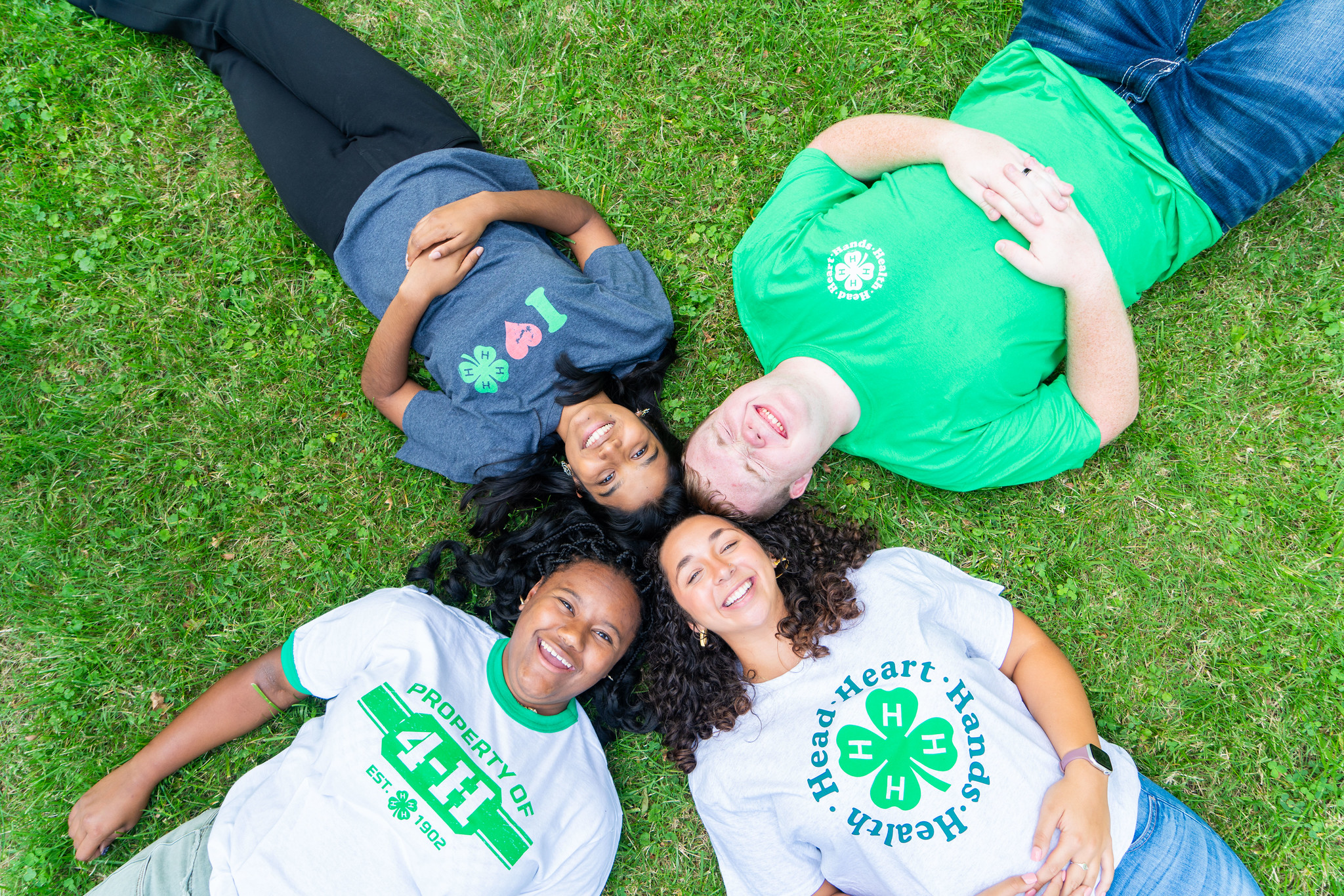 Four 4-H youth lie on the grass with their heads together in a circle, smiling up at the camera and wearing various 4-H T-shirts.