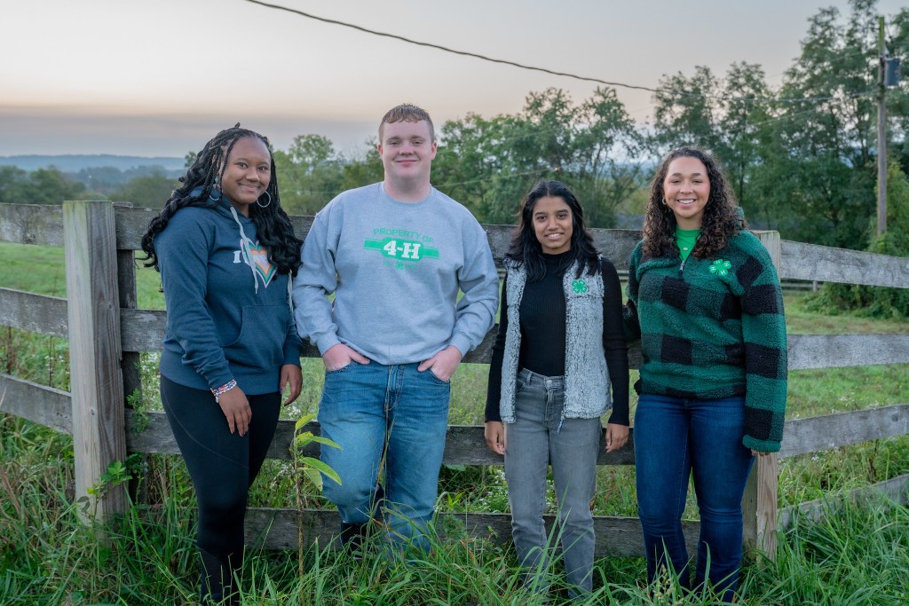 Four teenage 4-H members stand together and smile near a wooden fence with rolling hills in the background.