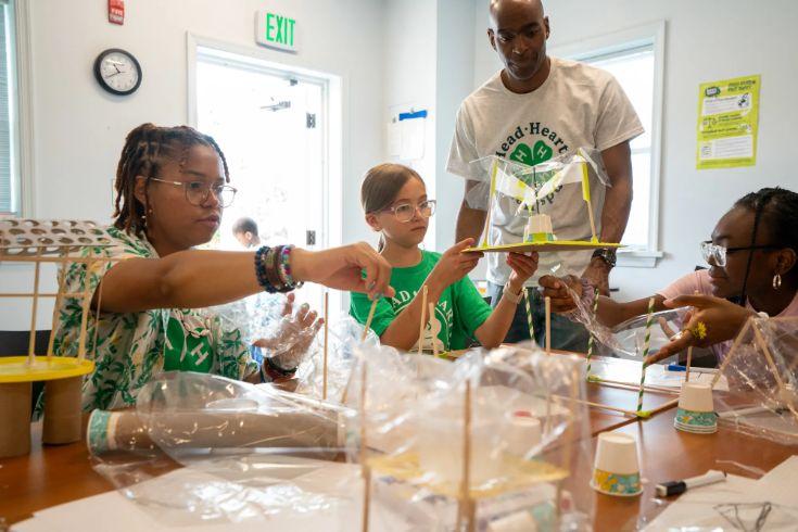 A group of children and a 4-H instructor work together on a hands-on STEM project at a table covered in craft supplies, building a model structure.