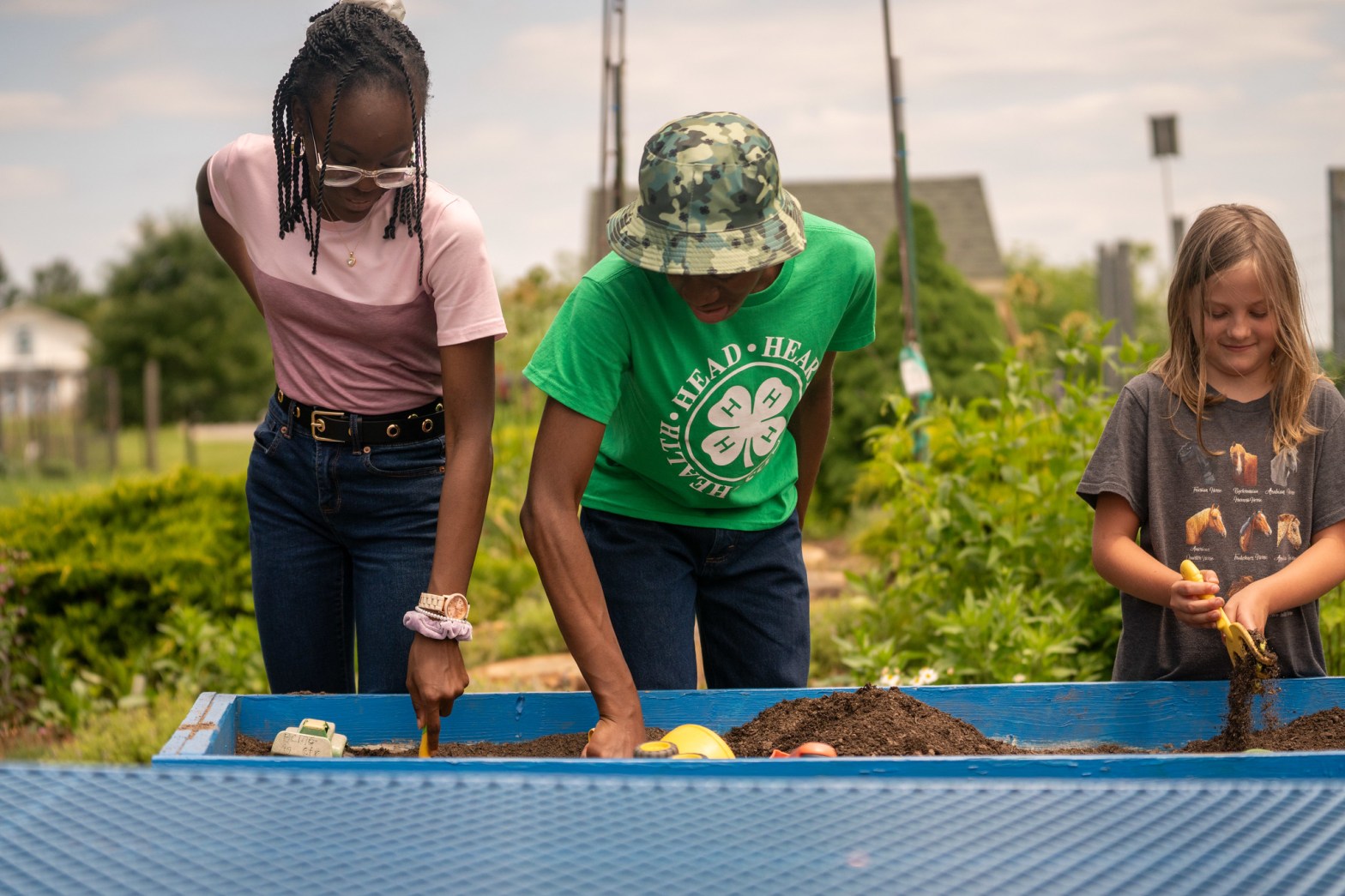 Three youth work together in a raised garden bed, digging in soil and using small tools.