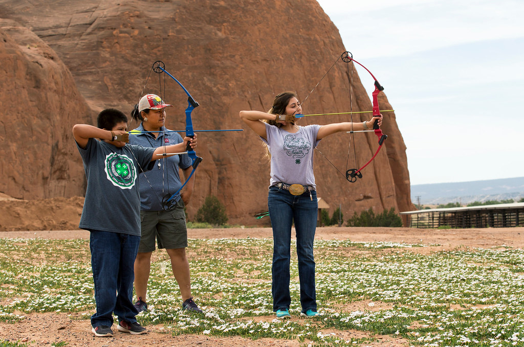 Three youth stand outdoors in a desert landscape practicing archery, each holding a drawn bow and aiming toward a target off camera. Large red rock formations rise in the background.