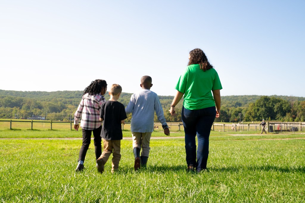 A 4-H leader walks through a grassy field with three children toward a wooden fence, with trees and hills in the background under a clear blue sky. They are seen from behind.