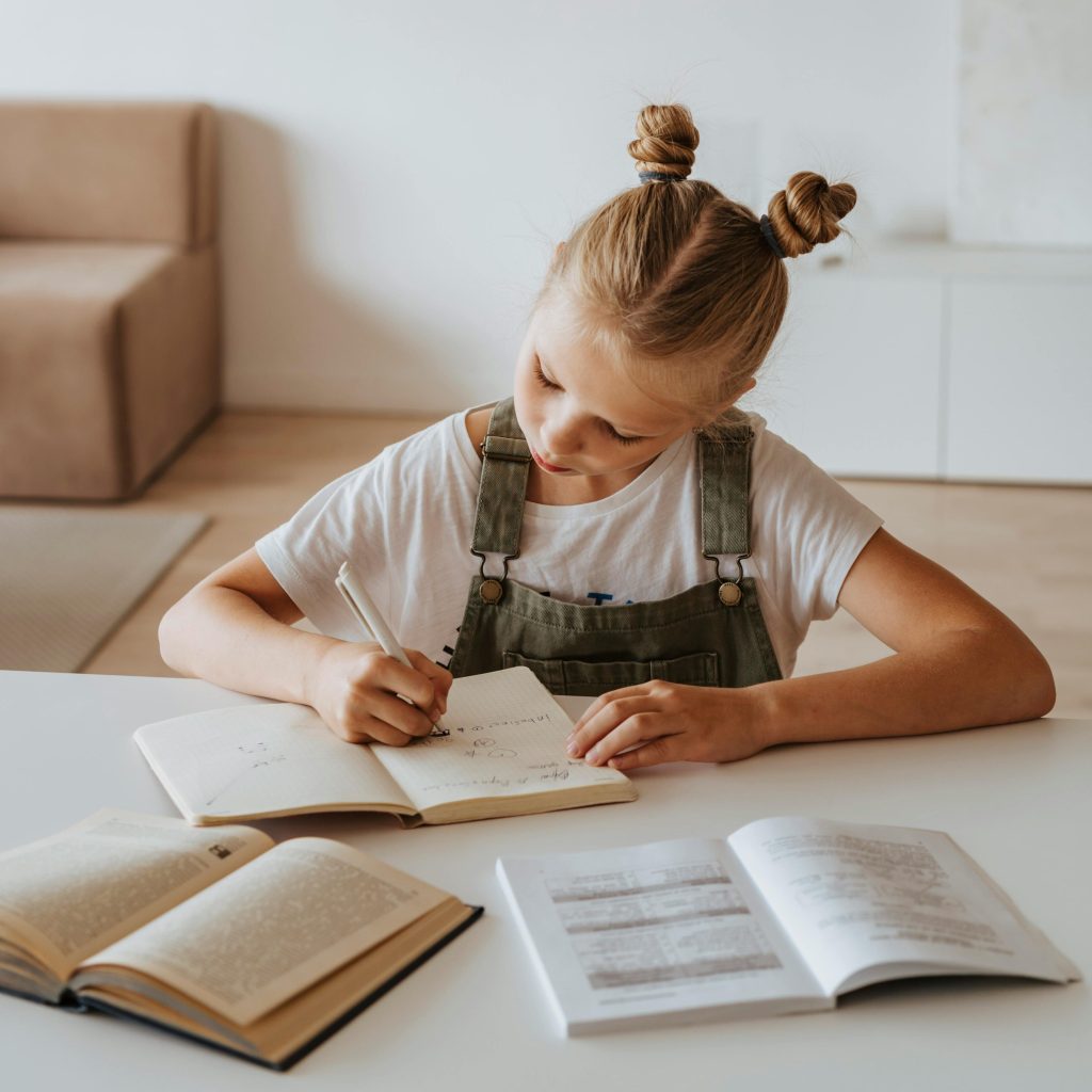 A young girl with blonde hair in two buns sits at a table, writing in a notebook. She is wearing a white shirt and green overalls, with open books spread out in front of her in a bright, modern room.