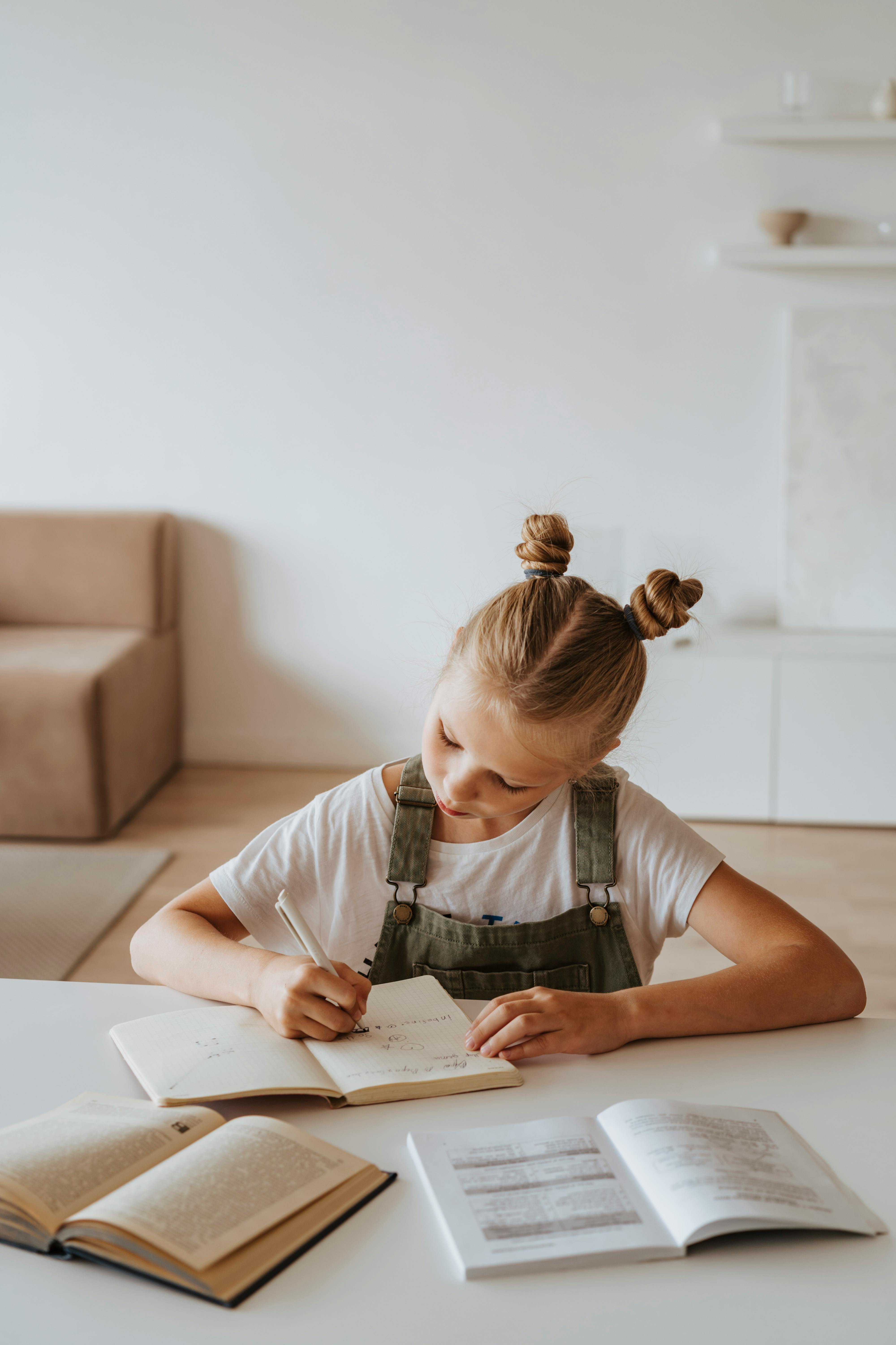 A young girl with blonde hair in two buns sits at a table, writing in a notebook. She is wearing a white shirt and green overalls, with open books spread out in front of her in a bright, modern room.