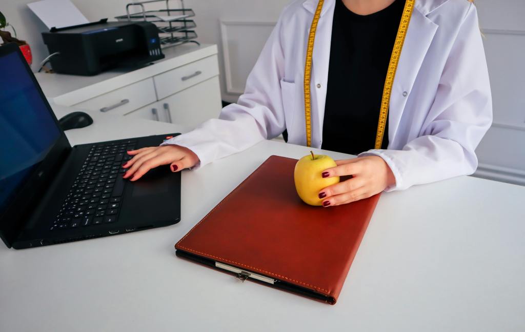Dietician typing in a laptop and holding an apple on a white table
