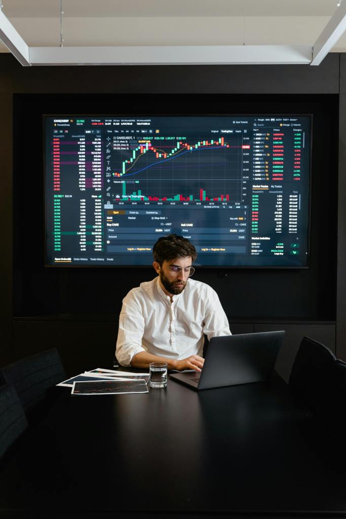 stock broker sitting in front of a large screen showing stats and trends with a black laptop on black table