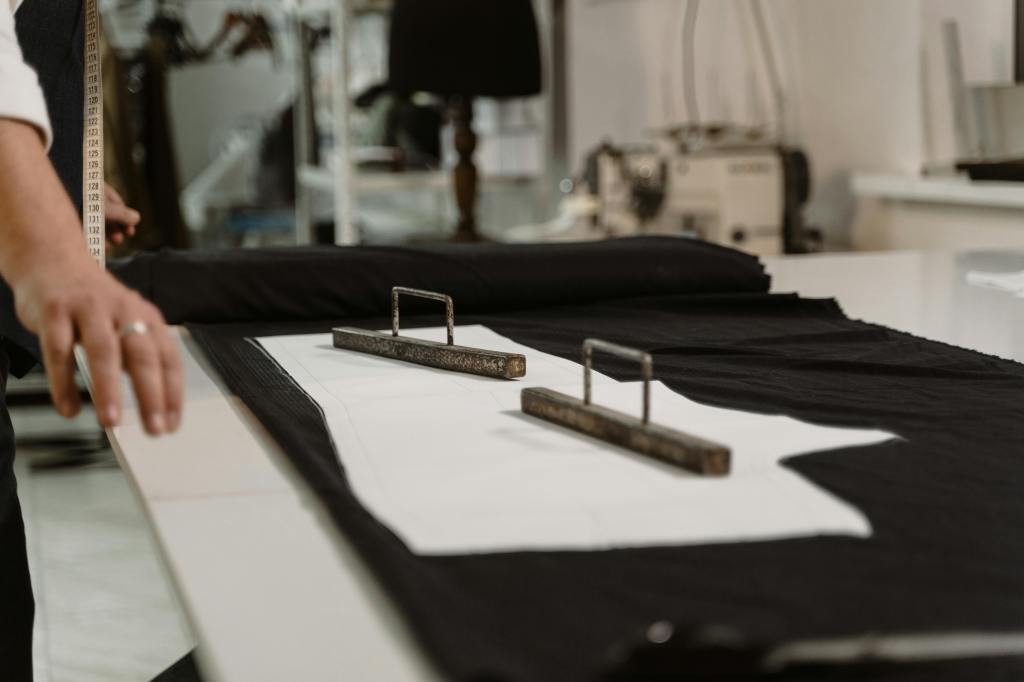 a close-up view of a pattern-making process in a tailoring workshop or atelier which shows a large piece of black fabric laid out on a white cutting table. White paper patterns are placed on top of the fabric. Two heavy, metal pattern weights with handles are used to hold the fabric and patterns securely in place