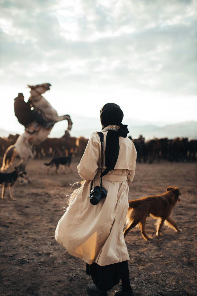a woman photographer wearing a head scarf with a camera slung over her shoulder and a man riding a horse which is rearing up on its hind legs in the background