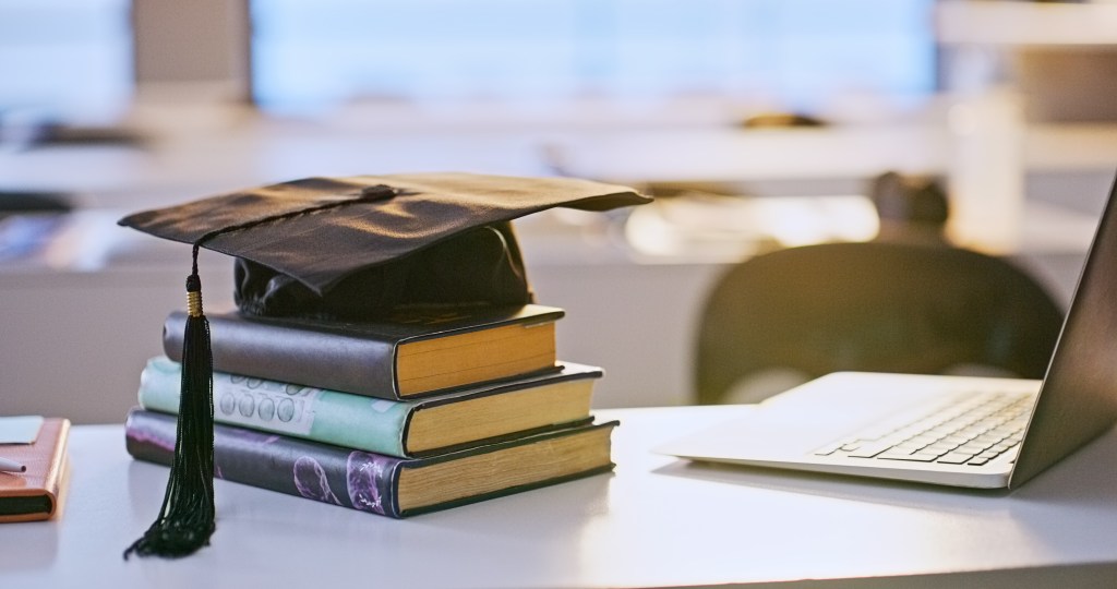A graduation cap rests on top of a stack of books beside an open laptop on a desk in a softly lit classroom.