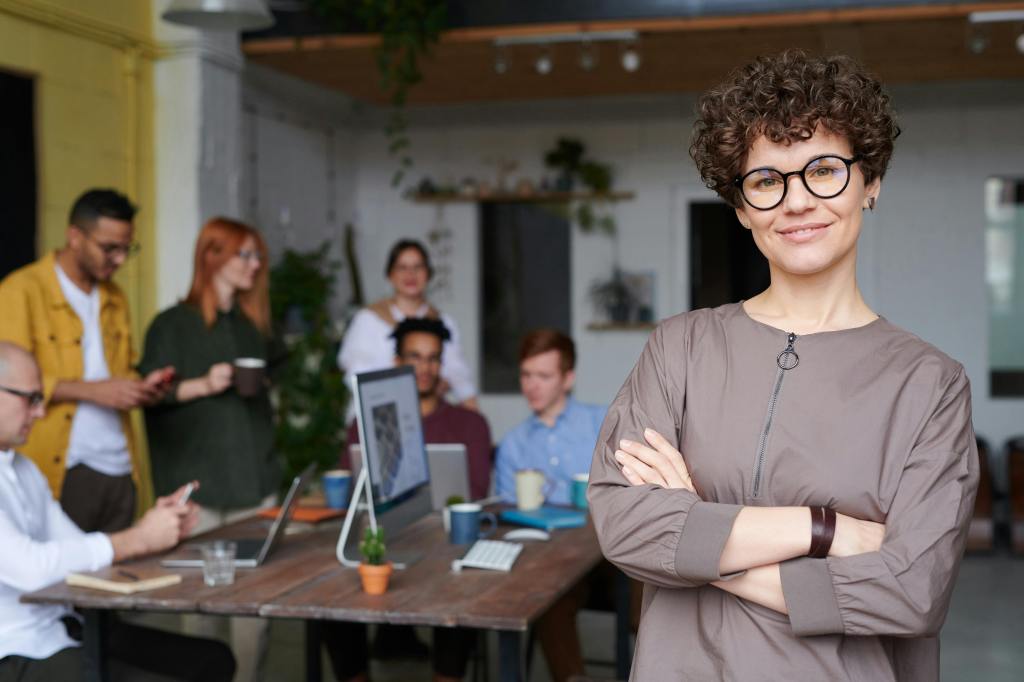 A woman with a short hair crossing her arms with random people sitting and standing behind her in an office or studio setting with a laptop on a table
