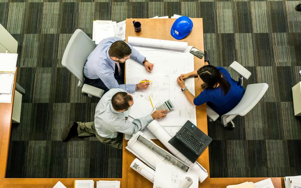 People sitting around a big wooden table with scrolls of paper, a phone, laptops, notes and plan communicating