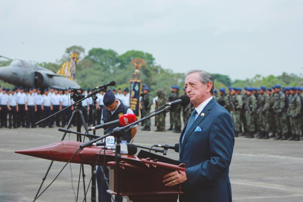 Older man in front of a podium with microphone with soldiers, pilots and a fighter jet in the background