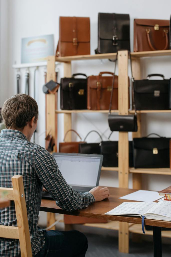 A guy sitting in front of laptop in shop with leather purses and bags on the shelves