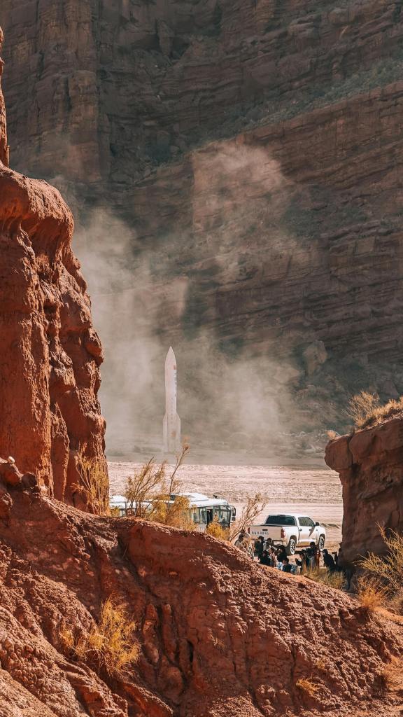 Group of people watching a rocket launch in a desert environment at the base of a canyon surrounded by red rocks