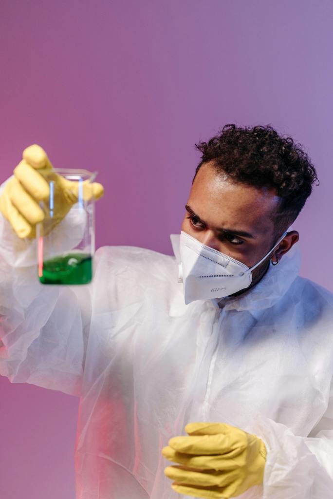 Scientist in a labcoat wearing a facemask looking at green liquid in a beaker