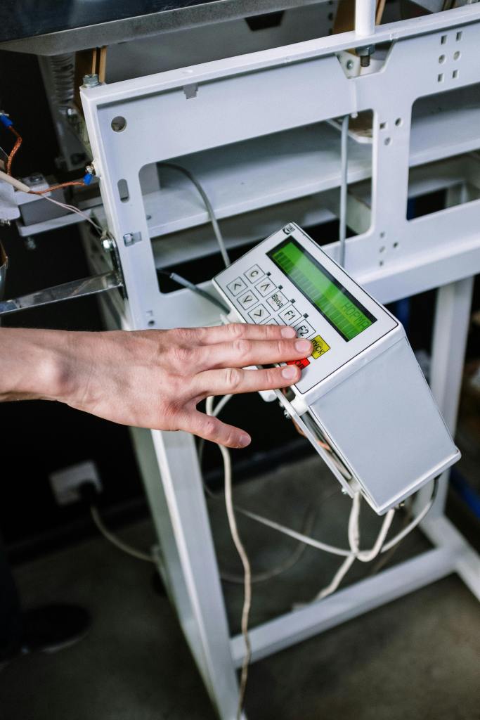 a close-up view of a person's hand operating an industrial control panel. The white, rectangular control panel has a green digital screen displaying text and data