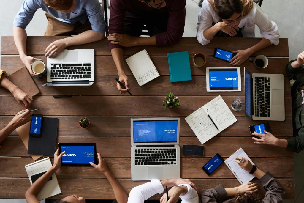 People sitting around a big wooden table with their phones, laptops and notes communicating