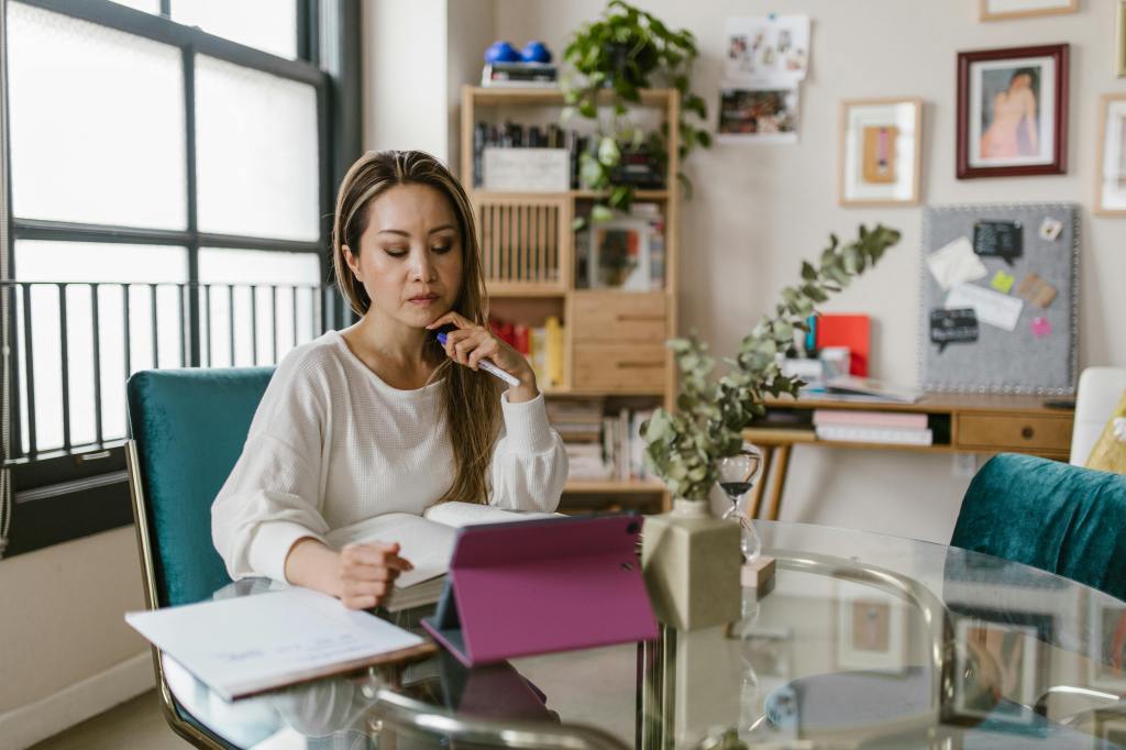Women wearing a white sweater working on a tablet on a round glass table with a notebook and a thick book open in front of her. seems to be a home or bright, casual office setting