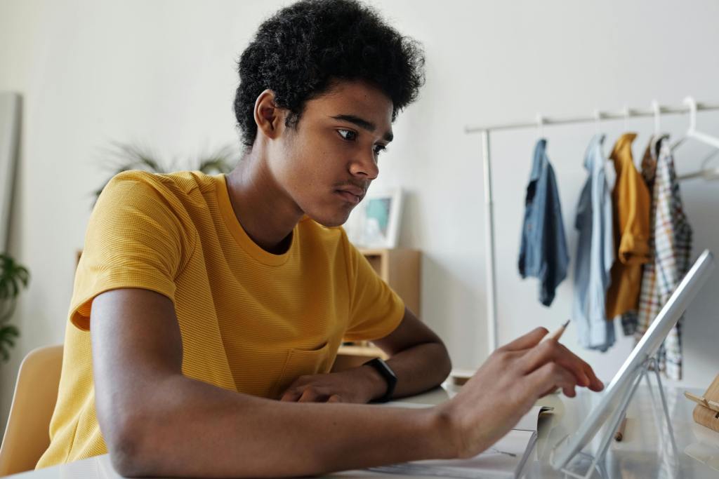 Young adult holding a pencil in his hand while using the touchscreen of his tablet, with clothes hanging on a rack in the background