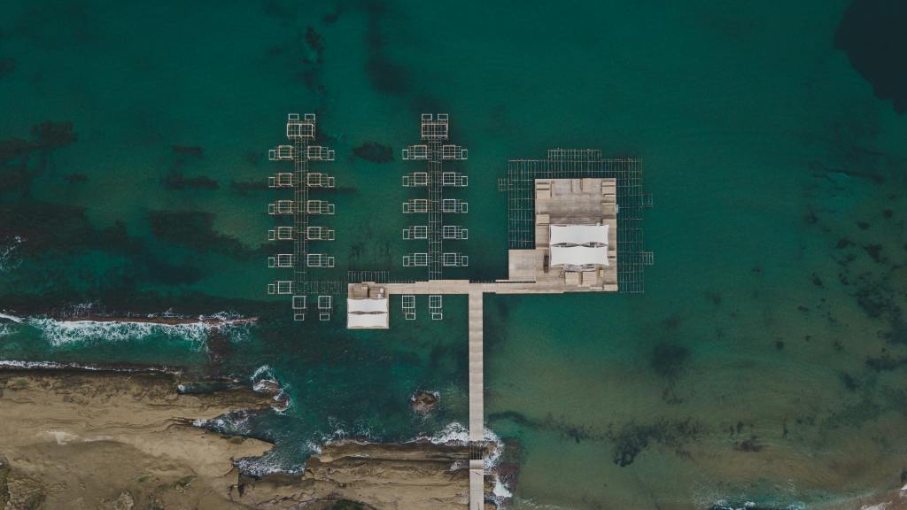 Bird's-eye view of a small coastal pier extending into turquoise ocean water. 