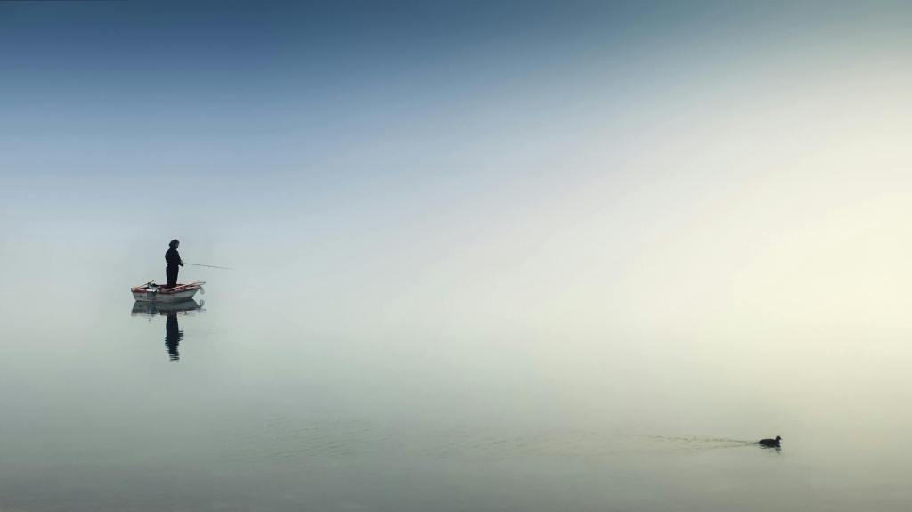Serene scene of a fisherman standing in a small boat on a calm, foggy lake with only a single duck in view