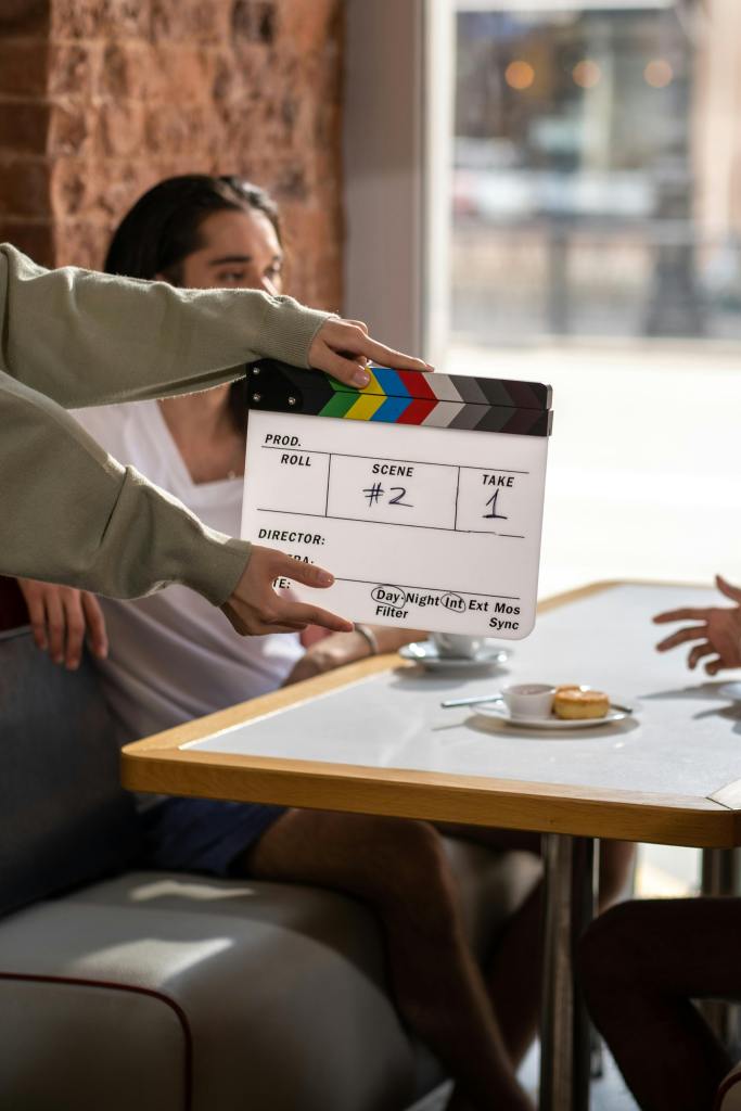 An indoor scene on a film set is depicted, with a person holding a clapperboard in the foreground. The clapperboard is positioned in front of another person seated at a table in what appears to be a cafe or restaurant setting