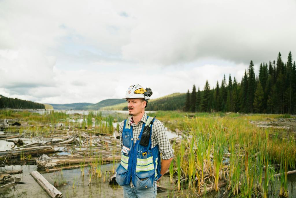 A man, presumably an Environmental educator, wearing professional safety gear stands in a marshy, natural environment.
