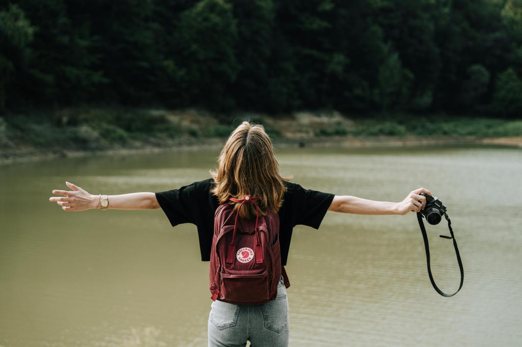 A female photographer with arms outstretched looking skyward wearing a backpack and holding a camera in her right hand standing next to a big river