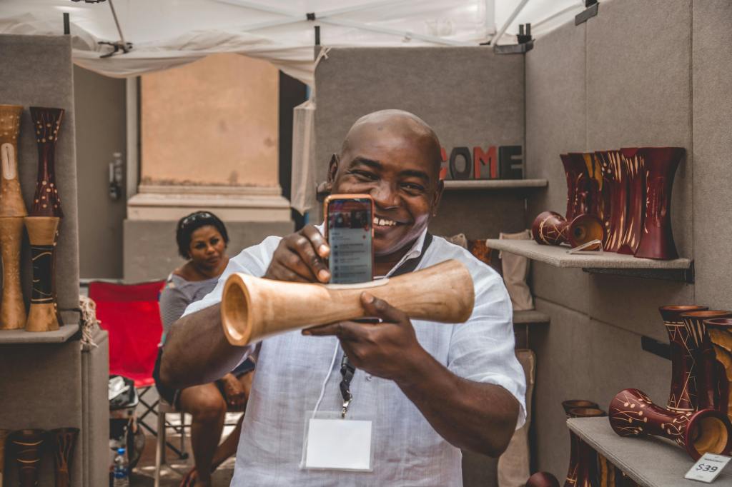 Shop owner showing a unique hand-carved, polished wooden decorative item that appears to be a mobile phone stand with a rich, reddish-brown finish