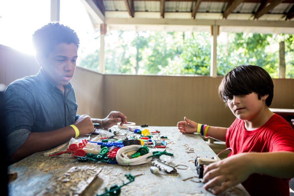 Two youth sit at a workshop table covered in craft and electronics materials, collaborating as they build a small wheeled project together. One adjusts parts while the other watches and assists.