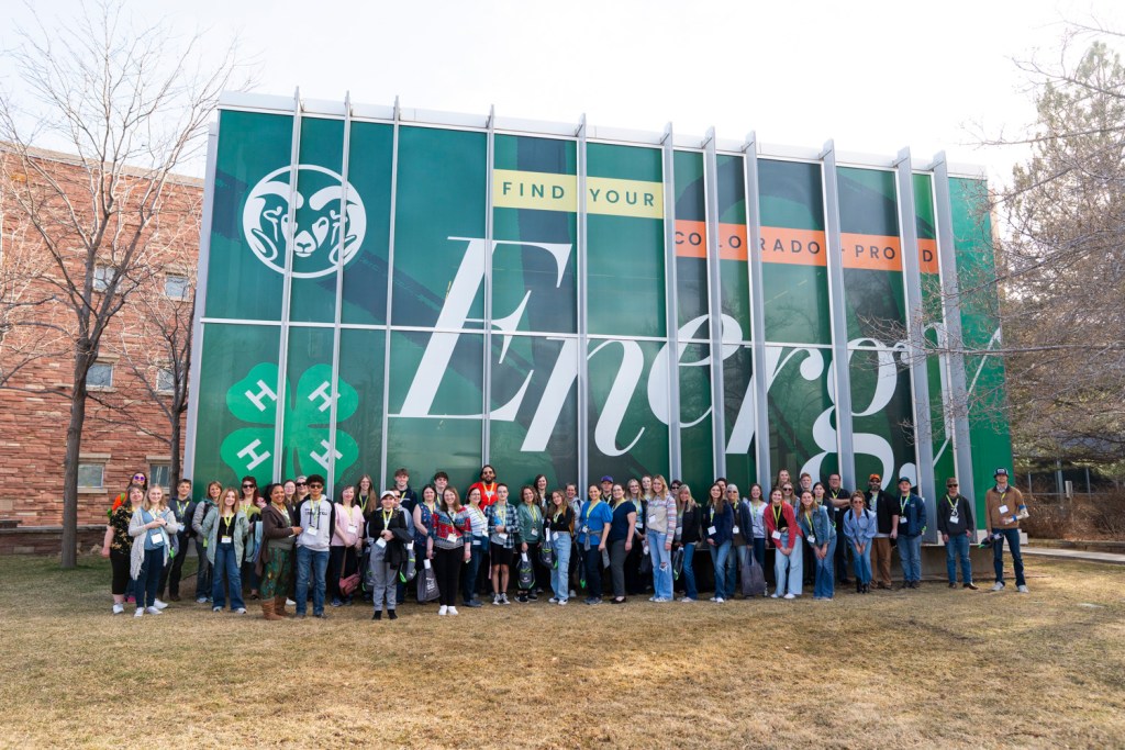 A large group of 4-H youth and adults stand outside in front of a tall green CSU building mural that reads ‘Find Your Energy,’ featuring the CSU Ram logo and a 4-H clover.