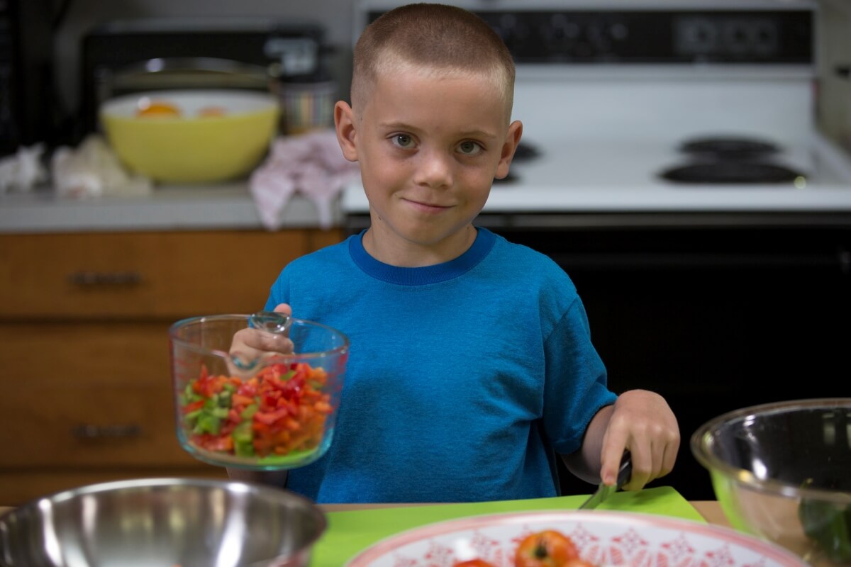 A young child smiles while standing in a kitchen, holding a measuring cup filled with chopped vegetables and a knife over a cutting board as part of a food preparation activity.