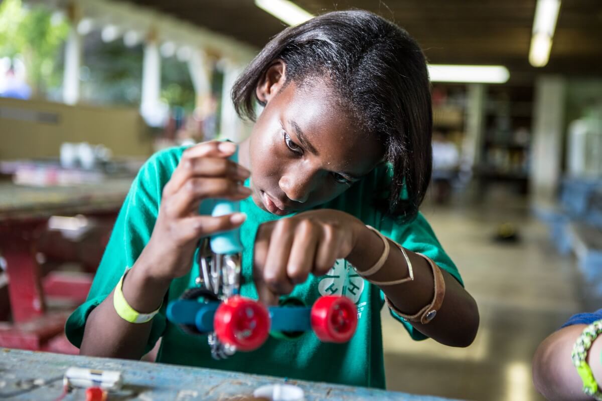A youth wearing a green 4-H shirt concentrates while using a hand tool to assemble or adjust a small wheeled project at a worktable indoors.