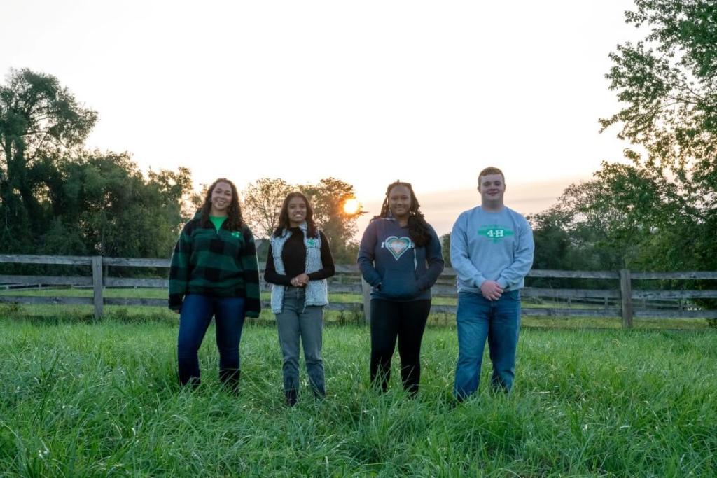 Four older 4-H youth stand in a grassy field at sunrise, with a wooden fence and trees behind them. They are smiling and wearing 4-H shirts and jackets.