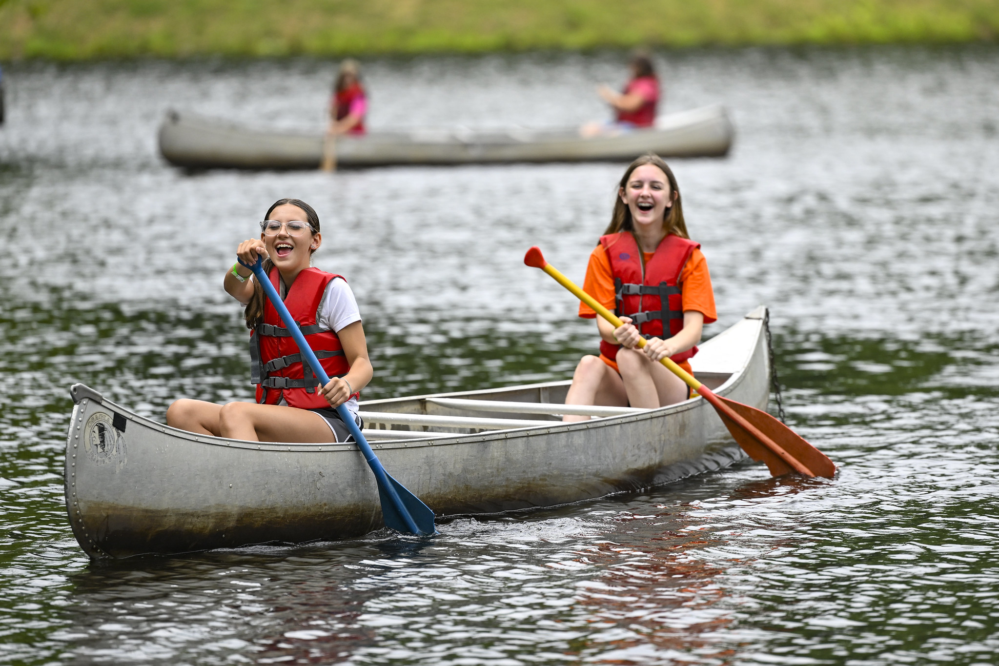 Two girls wearing life jackets paddle a canoe on a lake, smiling and laughing as they row together. Other youth in canoes paddle in the background. another two girls paddling a canoe blurred in the background.