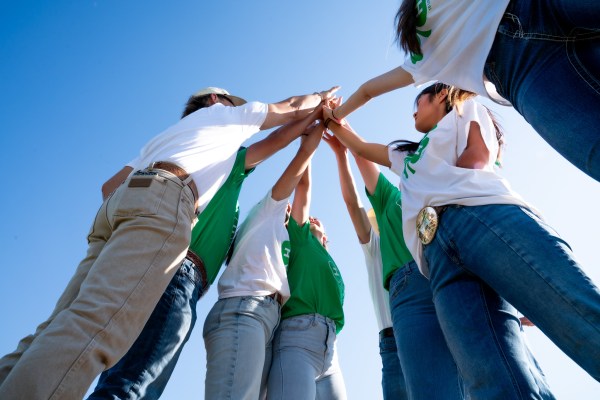 A group of 4-H youth standing in a circle outdoors, reaching their hands together in the center during a team huddle, photographed from a low angle against a clear blue sky.
