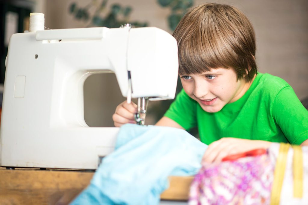 A child concentrates while sewing fabric on a sewing machine, guiding the material carefully with both hands.