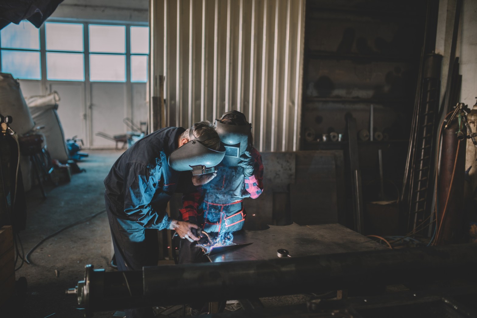 An adult and a youth wearing protective welding helmets work together on a metal welding project in a dimly lit workshop, with sparks visible from the welding torch.