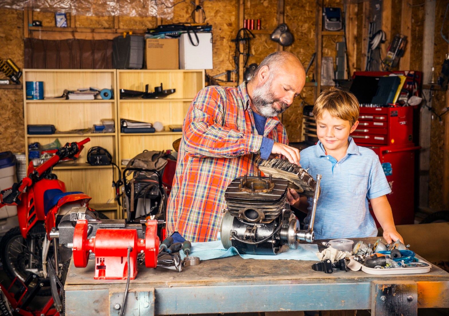 An adult and a young boy work together in a workshop, examining and assembling parts of a small engine on a workbench surrounded by tools and equipment.