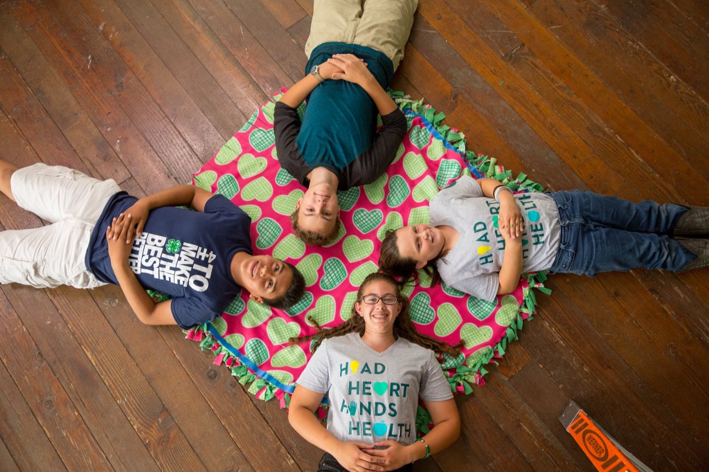 Four youth lie on a colorful heart-patterned blanket arranged in a circle on a wooden floor, smiling up at the camera while wearing 4-H themed shirts.