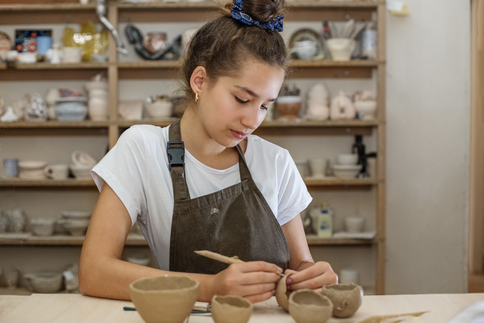 A young girl wearing an apron carefully shapes small clay bowls at a pottery table, with shelves of ceramic pieces and tools in the background.