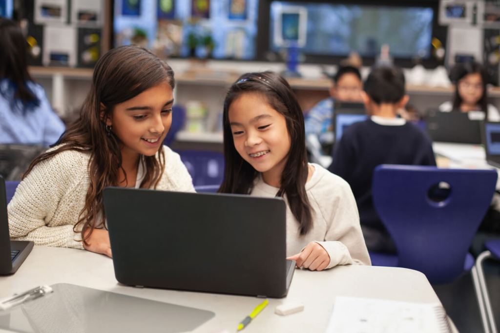 Two students sit side by side at a table in a classroom, smiling as they work together on a laptop, with other students using computers in the background.