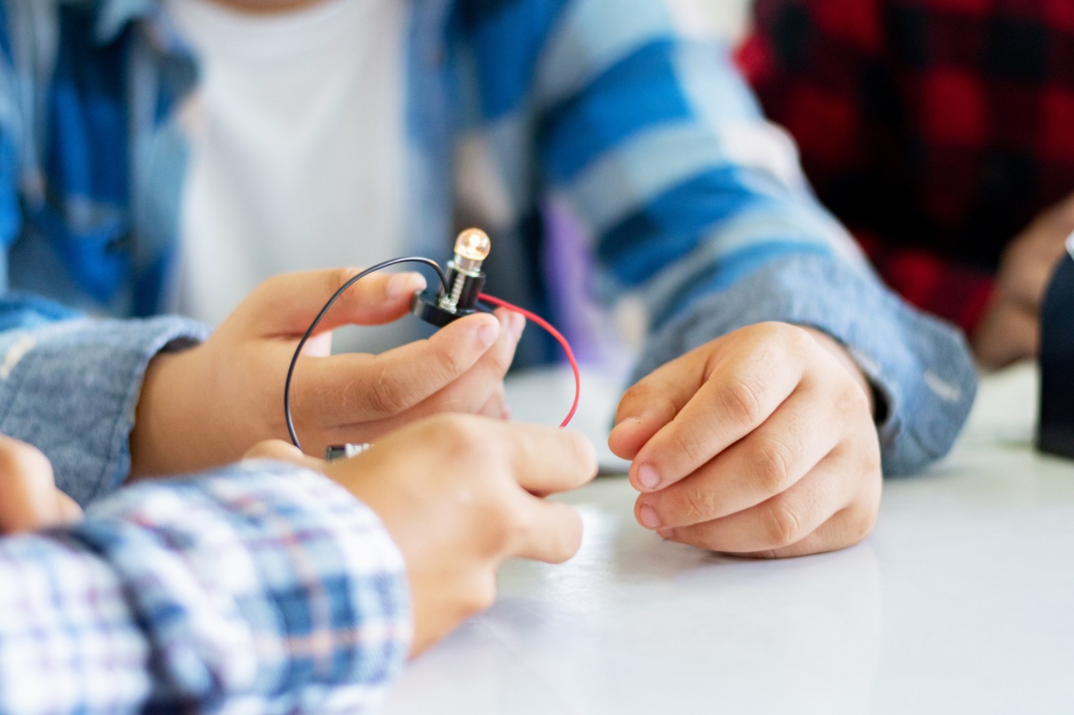 Close-up of children’s hands working together on a small electrical circuit, holding a battery-powered light bulb with red and black wires during a STEM activity.