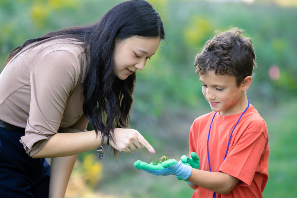 An adult and a young boy examine a small frog resting in the boy’s gloved hands during an outdoor nature activity.