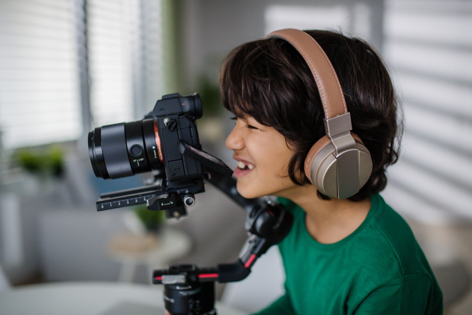 A child wearing large over-ear headphones smiles while looking through the viewfinder of a professional camera mounted on a stabilizer.