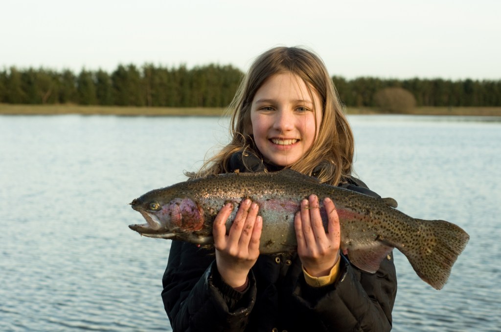 A smiling girl stands beside a lake holding a large rainbow trout with both hands after catching it outdoors.