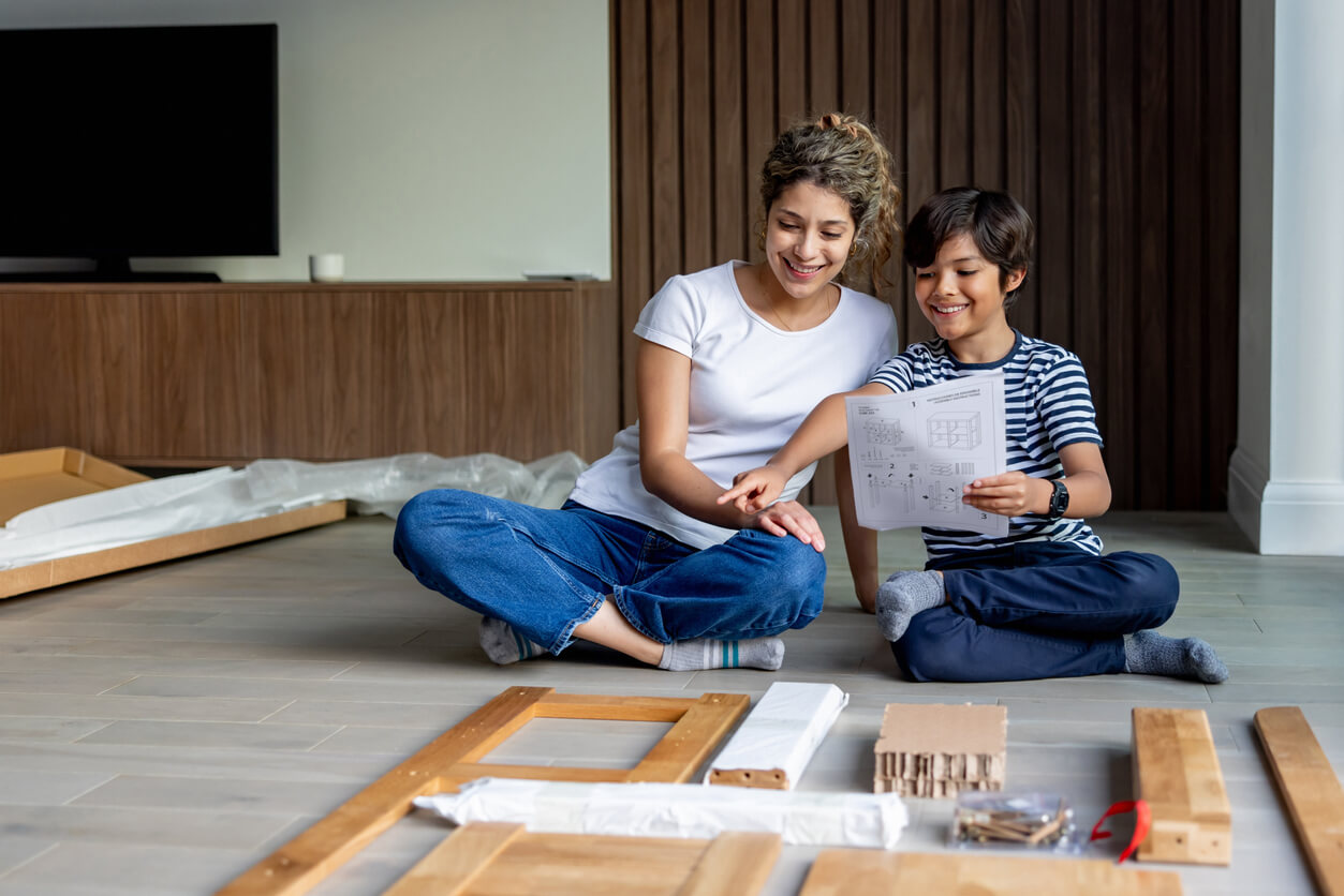 An adult and a child sit on the floor smiling while reviewing assembly instructions, with wooden furniture pieces and hardware spread out in front of them.