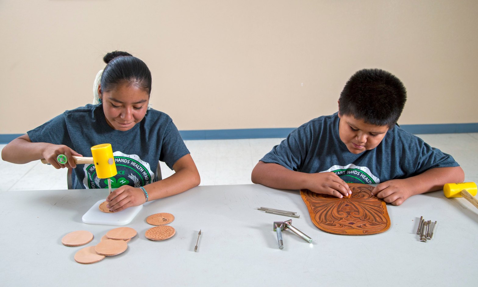 Two youth sit at a table working on leathercraft projects. One uses a yellow mallet and stamping tool to create designs on round leather pieces, while the other carefully carves patterns into a larger leather panel.
